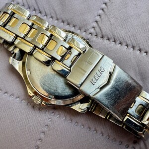 May include: A two-tone wristwatch featuring a silver and gold-colored metal link band. The clasp is engraved with the brand name RELIC. The watch is shown from the back, revealing a circular metal case back with a metallic finish and a crown on the side.