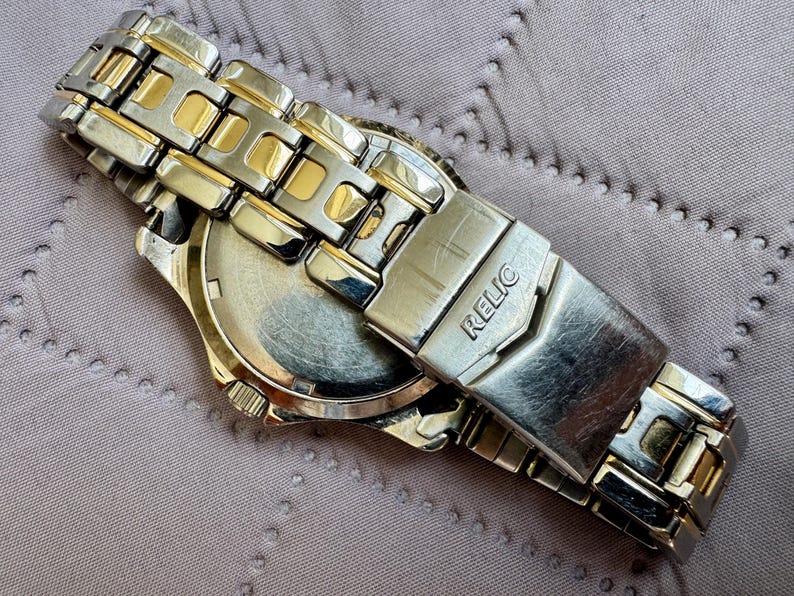 May include: A two-tone wristwatch featuring a silver and gold-colored metal link band. The clasp is engraved with the brand name RELIC. The watch is shown from the back, revealing a circular metal case back with a metallic finish and a crown on the side.