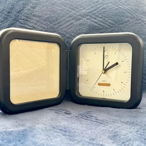 May include: A black, foldable travel clock with a white clock face and black hands. The clock is open, revealing a blank space for a photo. The clock face has black numbers and minute markers.