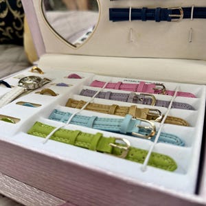 May include: A light pink jewelry box containing a watch with a white strap and multiple interchangeable watch bands in lime green, light blue, gold, lavender, and pink. The box features a heart-shaped mirror and slots for additional small accessories.