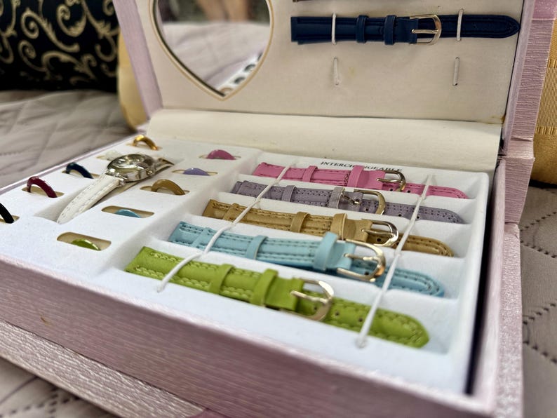 May include: A light pink jewelry box containing a watch with a white strap and multiple interchangeable watch bands in lime green, light blue, gold, lavender, and pink. The box features a heart-shaped mirror and slots for additional small accessories.