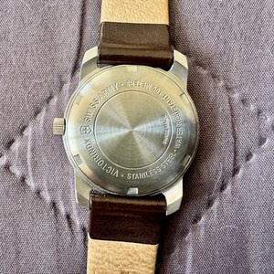 K&ouml;nnte beinhalten: Eine Victorinox Swiss Army Armbanduhr aus Edelstahl mit einem runden Geh&auml;useboden, der Gravuren wie Swiss Made und Water Resistant 100 Meters aufweist. Die Uhr ist an einem zweifarbigen Armband mit dunkelbraunem Leder und beigem Band befestigt.