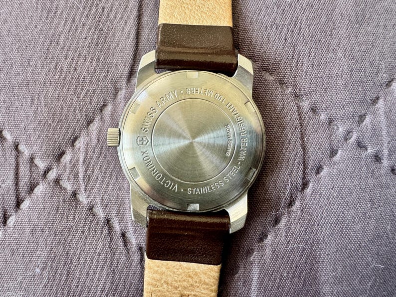 K&ouml;nnte beinhalten: Eine Victorinox Swiss Army Armbanduhr aus Edelstahl mit einem runden Geh&auml;useboden, der Gravuren wie Swiss Made und Water Resistant 100 Meters aufweist. Die Uhr ist an einem zweifarbigen Armband mit dunkelbraunem Leder und beigem Band befestigt.