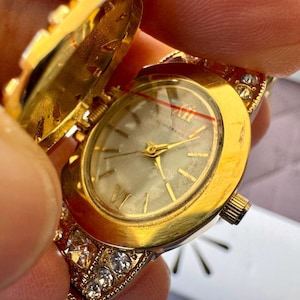 May include: A gold-toned locket watch with a hinged cover, revealing a watch face with Roman numerals. The watch is adorned with clear crystals and held open, showcasing the watch's intricate design.