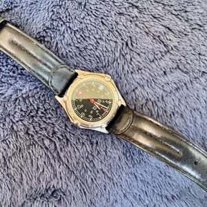 K&ouml;nnte beinhalten: Eine silberfarbene Armbanduhr mit schwarzem Zifferblatt und schwarzem Lederarmband. Das Zifferblatt hat wei&szlig;e Ziffern und einen roten Sekundenzeiger. Das Armband ist etwa 23 cm lang.