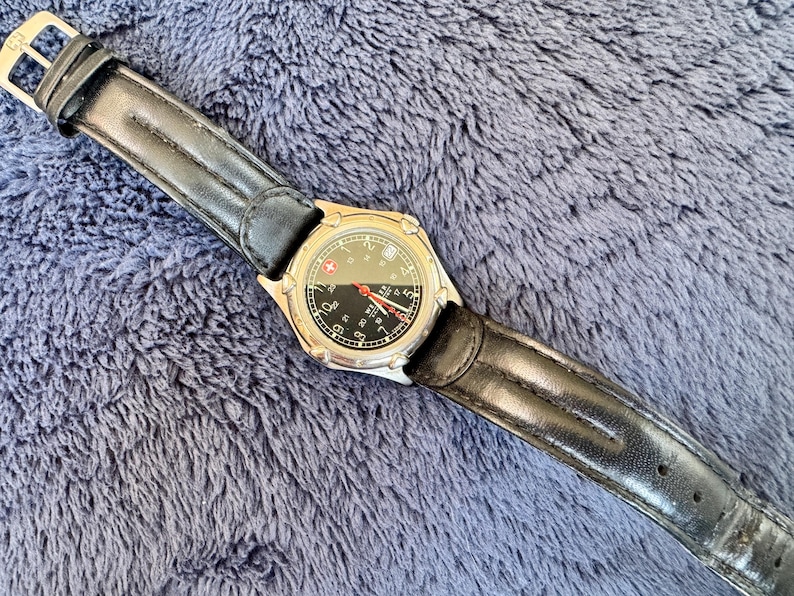 K&ouml;nnte beinhalten: Eine silberfarbene Armbanduhr mit schwarzem Zifferblatt und schwarzem Lederarmband. Das Zifferblatt hat wei&szlig;e Ziffern und einen roten Sekundenzeiger. Das Armband ist etwa 23 cm lang.
