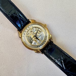 May include: A gold-toned Fossil watch with a detailed face featuring a figure and the word "AUTHENTIC". The watch has a black leather band with a textured, crocodile-like pattern.