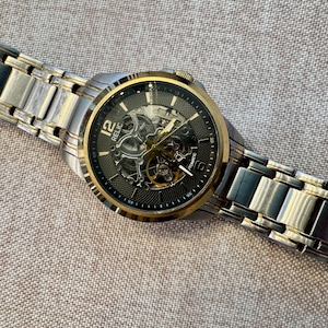 May include: A Relic automatic watch with a silver and gold-tone stainless steel band. The watch face is black with gold accents and visible internal mechanisms. The watch is displayed on a textured, neutral-toned surface.