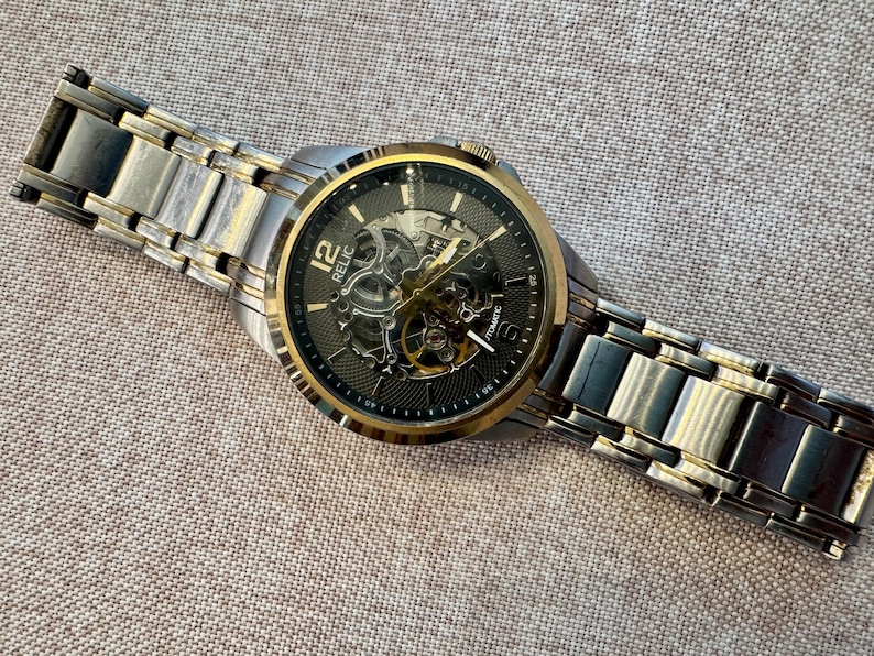 May include: A Relic automatic watch with a silver and gold-tone stainless steel band. The watch face is black with gold accents and visible internal mechanisms. The watch is displayed on a textured, neutral-toned surface.