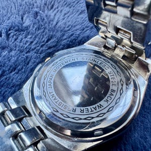 May include: Close-up of the back of a silver wristwatch. The watch back is stainless steel and features the text "Japan Movement", "Water Resistant", and "Stainless Steel Back". The watch band is also silver.