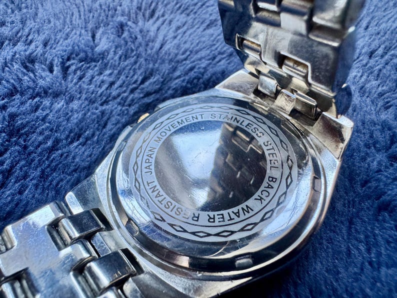 May include: Close-up of the back of a silver wristwatch. The watch back is stainless steel and features the text "Japan Movement", "Water Resistant", and "Stainless Steel Back". The watch band is also silver.