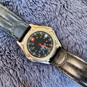 K&ouml;nnte beinhalten: Eine silberfarbene Armbanduhr mit schwarzem Zifferblatt und schwarzem Lederarmband. Das Zifferblatt hat wei&szlig;e Ziffern, einen roten Sekundenzeiger und ein Datumsfenster. Das Armband ist aus schwarzem Leder. Die Uhr hat ein Schweizer Kreuz-Logo.