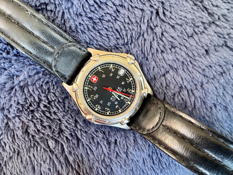 K&ouml;nnte beinhalten: Eine silberfarbene Armbanduhr mit schwarzem Zifferblatt und schwarzem Lederarmband. Das Zifferblatt hat wei&szlig;e Ziffern, einen roten Sekundenzeiger und ein Datumsfenster. Das Armband ist aus schwarzem Leder. Die Uhr hat ein Schweizer Kreuz-Logo.