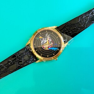 Peut inclure: Une montre dor&eacute;e avec un cadran noir repr&eacute;sentant des personnages de dessins anim&eacute;s. La montre a un bracelet en cuir noir avec un motif crocodile. La montre est pos&eacute;e sur un fond turquoise.