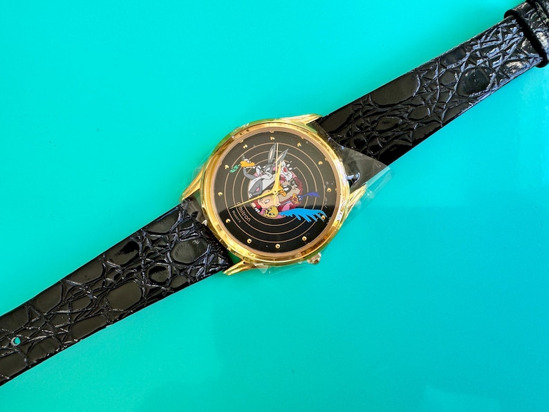 Peut inclure: Une montre dor&eacute;e avec un cadran noir repr&eacute;sentant des personnages de dessins anim&eacute;s. La montre a un bracelet en cuir noir avec un motif crocodile. La montre est pos&eacute;e sur un fond turquoise.