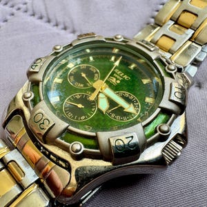 May include: A Relic Wet wristwatch featuring a round green textured dial with three sub-dials, luminous hands, and a silver-tone bezel with engraved numbers. The watch has a two-tone stainless steel link bracelet in silver and gold tones.