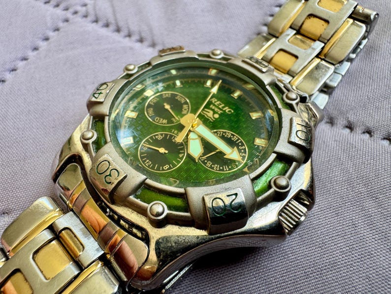 May include: A Relic Wet wristwatch featuring a round green textured dial with three sub-dials, luminous hands, and a silver-tone bezel with engraved numbers. The watch has a two-tone stainless steel link bracelet in silver and gold tones.