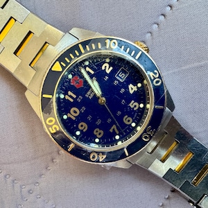May include: A Swiss Army wristwatch featuring a deep blue dial with white Arabic numerals, a date window, and a rotating blue bezel with yellow markings. The watch has a two-tone silver and gold-colored metal link bracelet with a secure clasp.