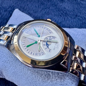 May include: A silver and gold-toned wristwatch with a white face and green hands. The watch features a silver and gold band and is marked "Paul Jar" and "Quartz". The watch is resting on a light blue surface.