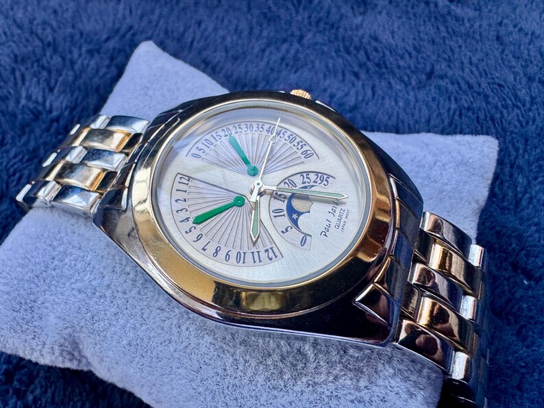 May include: A silver and gold-toned wristwatch with a white face and green hands. The watch features a silver and gold band and is marked "Paul Jar" and "Quartz". The watch is resting on a light blue surface.