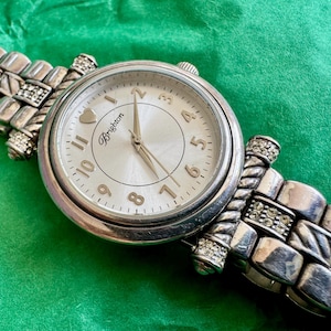 May include: A silver-tone analog wristwatch with a round face, featuring a white dial with gold-tone Arabic numerals, a heart icon at the twelve o'clock position, and the Brighton logo. The metal link band includes twisted accents and clear crystals.