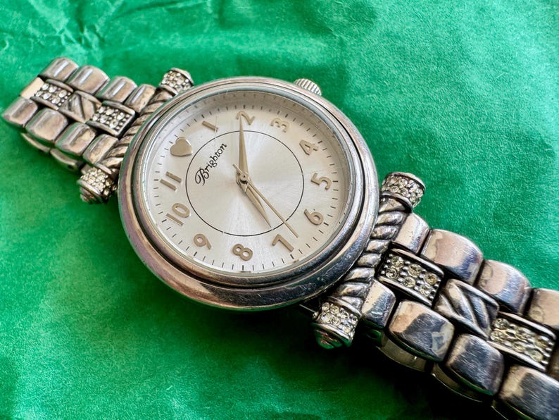 May include: A silver-tone analog wristwatch with a round face, featuring a white dial with gold-tone Arabic numerals, a heart icon at the twelve o'clock position, and the Brighton logo. The metal link band includes twisted accents and clear crystals.