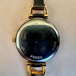 May include: A Fossil brand wristwatch, featuring a black leather strap and gold-toned accents. The watch face is round with a dark, reflective surface. The back of the watch is visible, displaying the text "ALL STAINLESS STEEL", "5 ATM", "ES3111", and "251210".