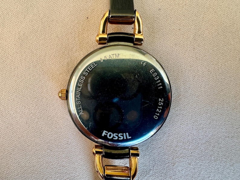 May include: A Fossil brand wristwatch, featuring a black leather strap and gold-toned accents. The watch face is round with a dark, reflective surface. The back of the watch is visible, displaying the text "ALL STAINLESS STEEL", "5 ATM", "ES3111", and "251210".