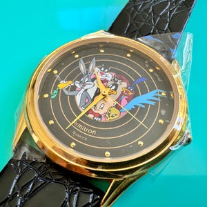 Peut inclure: Montre Armitron Quartz dor&eacute;e avec un cadran noir repr&eacute;sentant les personnages de Looney Tunes. La montre a un bracelet en cuir noir et est neuve dans son emballage d'origine.