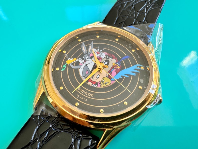 Peut inclure: Montre Armitron Quartz dor&eacute;e avec un cadran noir repr&eacute;sentant les personnages de Looney Tunes. La montre a un bracelet en cuir noir et est neuve dans son emballage d'origine.