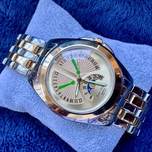 May include: A gold and silver-toned wristwatch with a light-colored face. The watch features a unique dial design with multiple hands and a date display. The watch band is a combination of silver and gold links.