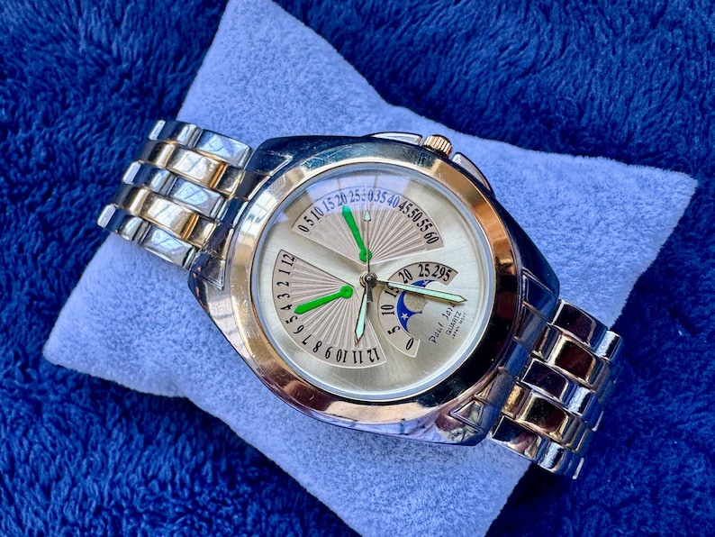 May include: A gold and silver-toned wristwatch with a light-colored face. The watch features a unique dial design with multiple hands and a date display. The watch band is a combination of silver and gold links.