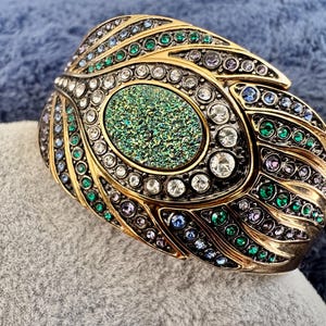 May include: A gold-toned cuff bracelet with an oval, textured, green center stone. The bracelet is adorned with clear, green, blue, and purple gemstones. The design features a feather-like pattern, creating a luxurious and ornate accessory.