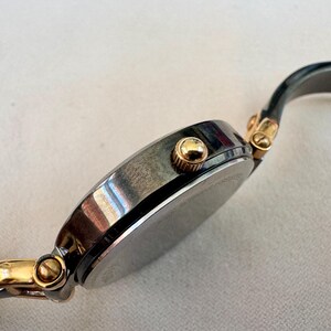 May include: A silver-toned wristwatch with gold accents. The watch has a round face with a dark dial and a gold crown. The band is a dark gray color. The watch is a classic style.