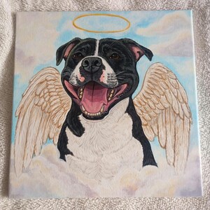 Può includere: Opera d'arte su tela quadrata raffigurante un cane bianco e nero sorridente con ali d'angelo e un'aureola dorata. Il cane è dipinto su uno sfondo di nuvole e cielo blu. La bocca del cane è aperta, rivelando la sua lingua rosa.