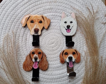 Puede incluir: Cuatro ganchos en forma de cabeza de perro con caras pintadas. Los ganchos son de madera y tienen un gancho de metal negro o plateado. Los perros son un golden retriever, un perro blanco, un spaniel marrón y blanco y un beagle.