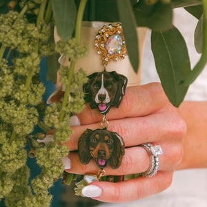 May include: A decorative charm accessory featuring a gold-toned filigree brooch with an iridescent center stone, connected to two hand-painted dog portraits. The top dog is brown and white, and the bottom dog is solid brown, both with pink tongues.