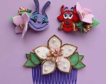May include: Three felt hair clips with a flower design. The first clip features a purple bug with a pink flower. The second clip features a red dragon with a pink flower. The third clip features a white flower with gold accents and a blue comb.