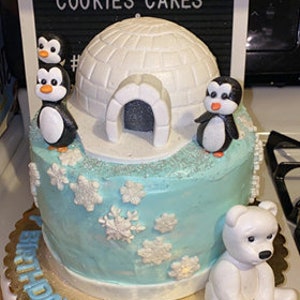 May include: A two-tiered blue and white birthday cake decorated with a white igloo, penguins, and snowflakes. The cake is on a gold-rimmed cake board with the name "Derek" written in gold icing. The cake is topped with a white polar bear figurine.