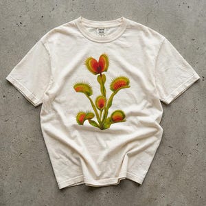 May include: A cream-coloured t-shirt with a colourful illustration of a Venus flytrap plant. The graphic shows the plant's distinctive red and green traps and stems. The shirt has a classic crew neck and short sleeves.