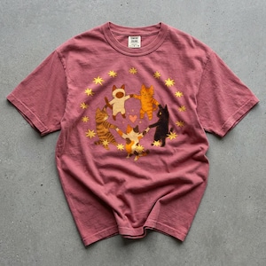 May include: A dusty rose-colored t-shirt featuring a graphic of five cats holding hands in a circle, surrounded by yellow stars. The cats are in various colors, including orange, black, and calico. The shirt has a crew neck and short sleeves.
