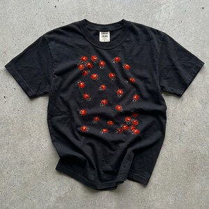 Può includere: Una t-shirt nera a maniche corte con un motivo sparso di piccole coccinelle rosse sul davanti. La maglia presenta uno scollo rotondo e un'etichetta interna bianca sul colletto con il testo Comfort Colors visibile al centro.