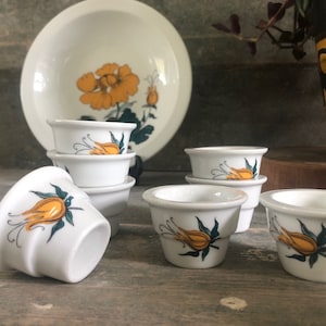 May include: Set of six white ceramic egg cups with yellow and green floral designs. The cups are stacked in pairs and feature a single yellow flower with green leaves on each cup.