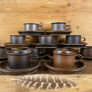 May include: A set of ten brown and black ceramic coffee cups and saucers. The cups and saucers are stacked in a pyramid shape on a wooden surface.