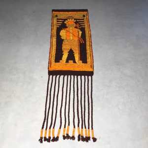 May include: A brown and orange woven wall hanging with a geometric design of a person holding a stick. The hanging has a fringe of brown yarn with orange accents.
