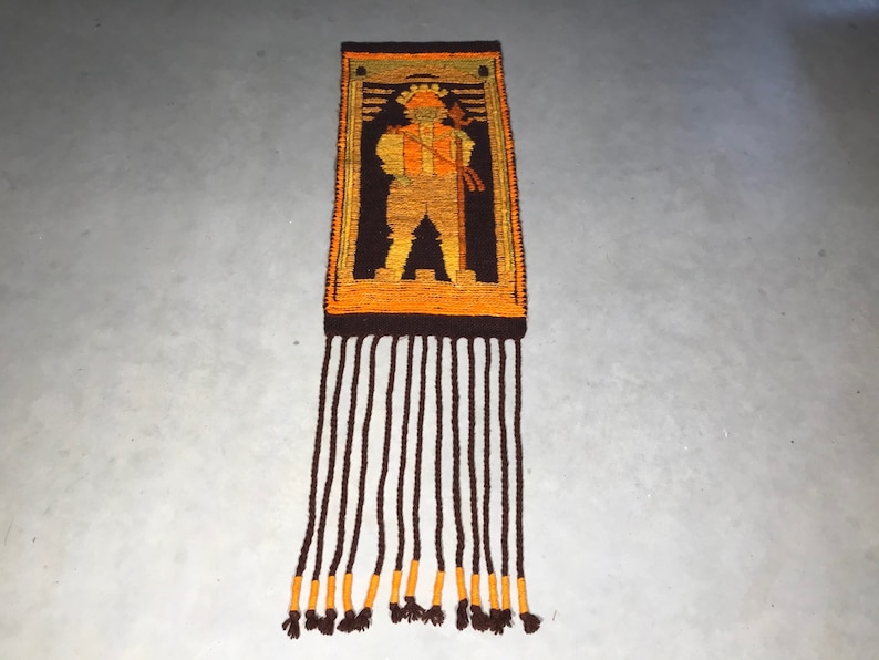 May include: A brown and orange woven wall hanging with a geometric design of a person holding a stick. The hanging has a fringe of brown yarn with orange accents.