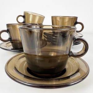 K&ouml;nnte beinhalten: Set aus vier braunen Glas-Teetassen und Untertassen. Die Tassen und Untertassen sind dunkelbraun und haben eine glatte, abgerundete Form.