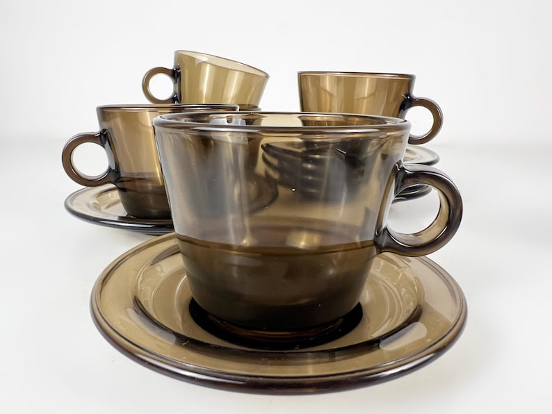 K&ouml;nnte beinhalten: Set aus vier braunen Glas-Teetassen und Untertassen. Die Tassen und Untertassen sind dunkelbraun und haben eine glatte, abgerundete Form.