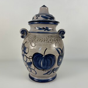 May include: A ceramic Rumtopf pot with a lid, featuring a light grey base and dark blue accents. The pot is adorned with a blue apple design and the word "Rumtopf" is embossed on the front. It has two small handles.