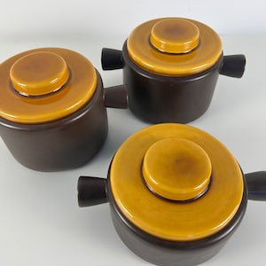 Puede incluir: Conjunto de tres cazuelas de cerámica marrón con tapas amarillas. Las tapas tienen un círculo central elevado.